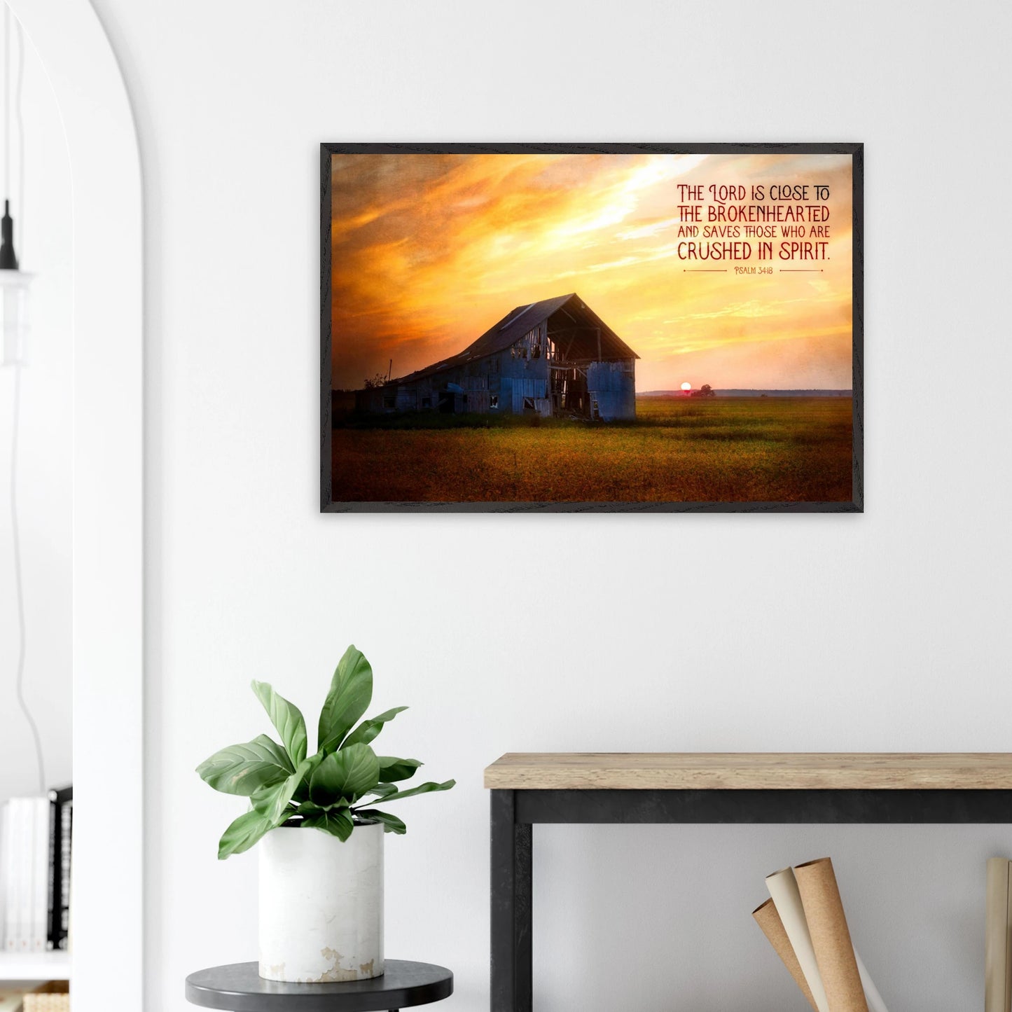 Framed artwork of a sunset over a barn with a motivational quote on a white wall above a desk.