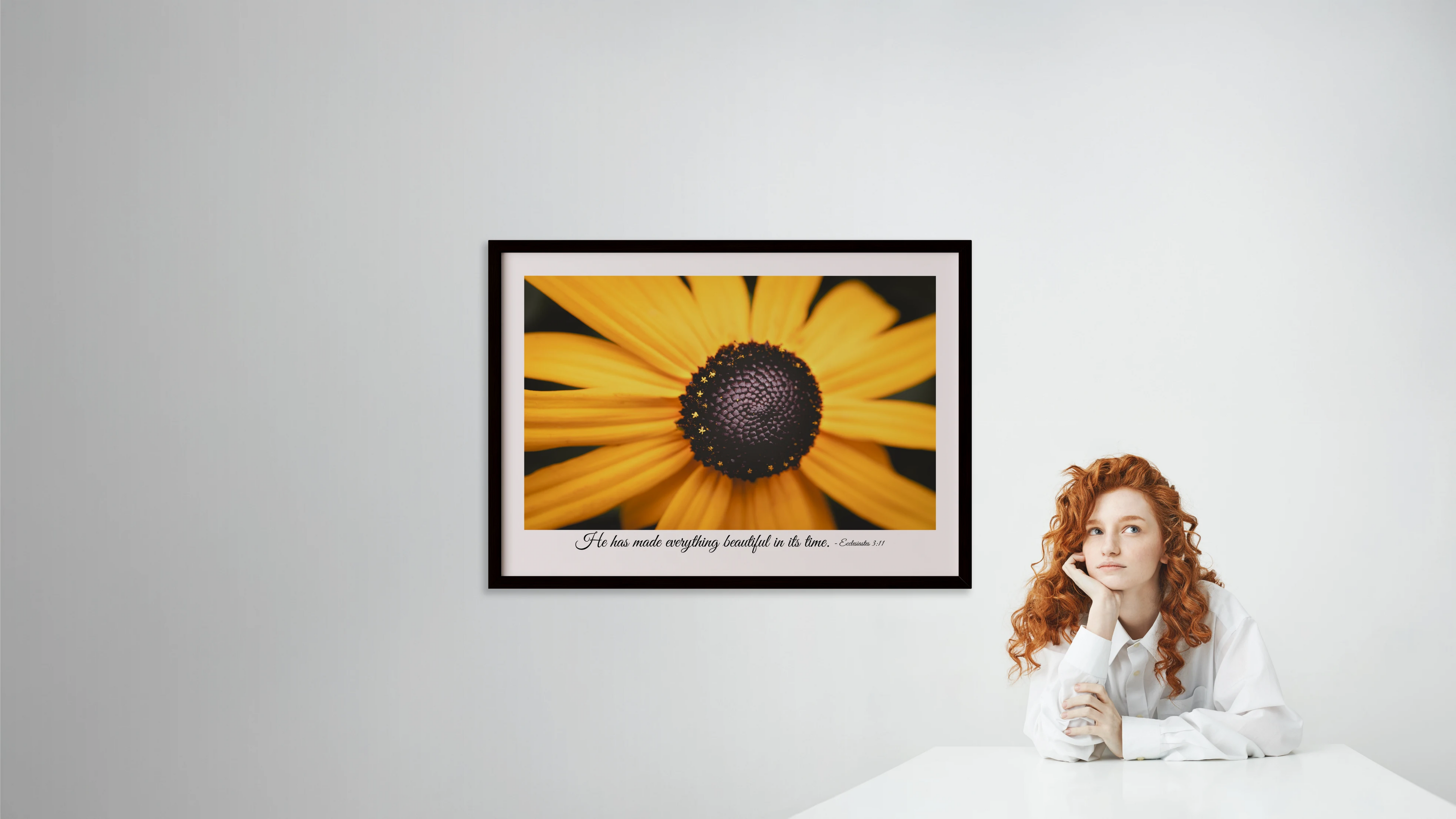 Woman sitting next to a framed picture of a sunflower on a white wall.