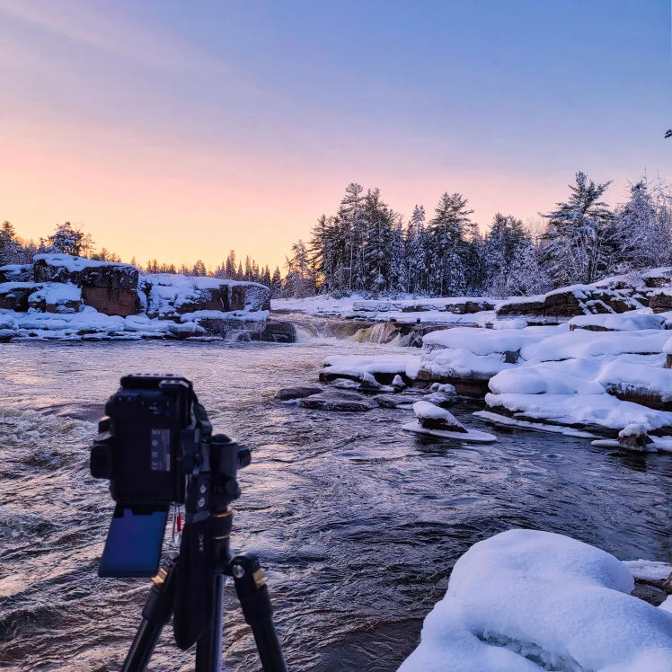 Camera on a tripod capturing a snowy landscape with a river and trees at sunset.