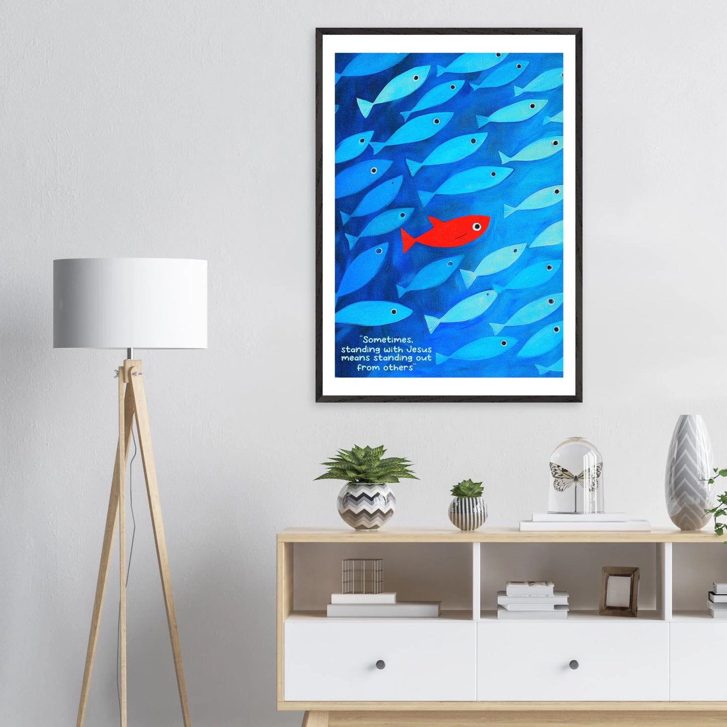 Framed wall art featuring a school of blue fish with a single red fish, and text that reads: 'Sometimes standing with Jesus means standing out from others.' displayed in a bright modern minimalist interior