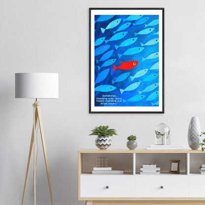 Framed wall art featuring a school of blue fish with a single red fish, and text that reads: 'Sometimes standing with Jesus means standing out from others.' displayed in a bright modern minimalist interior