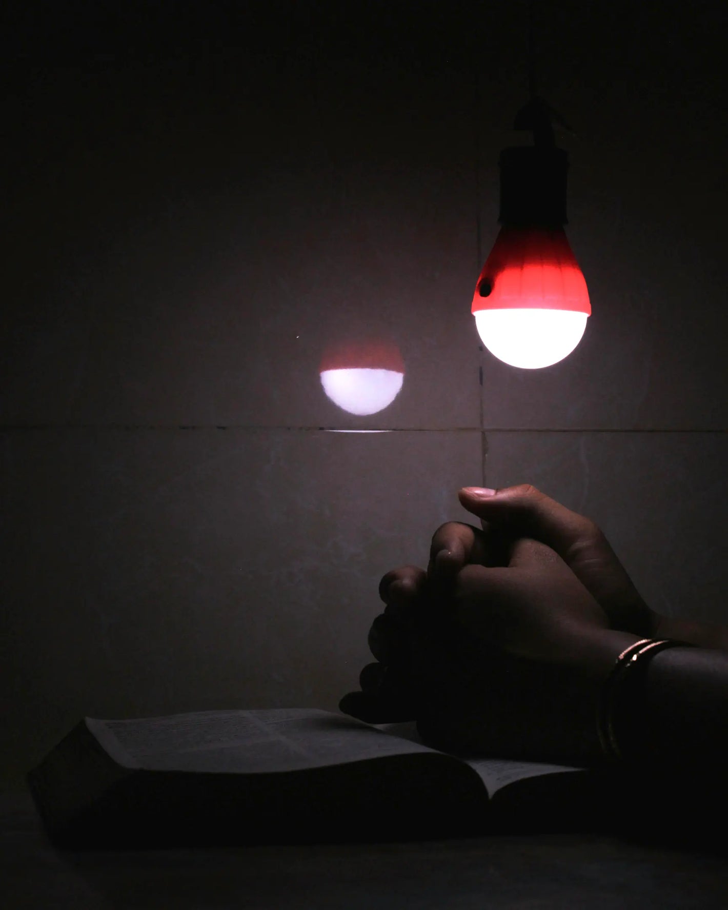 Two light bulbs, one red and one white, hanging on a dark wall with a hand reaching towards them.