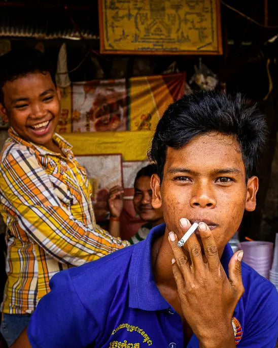 Two young men, one smoking a cigarette, in a casual setting with posters on the wall.