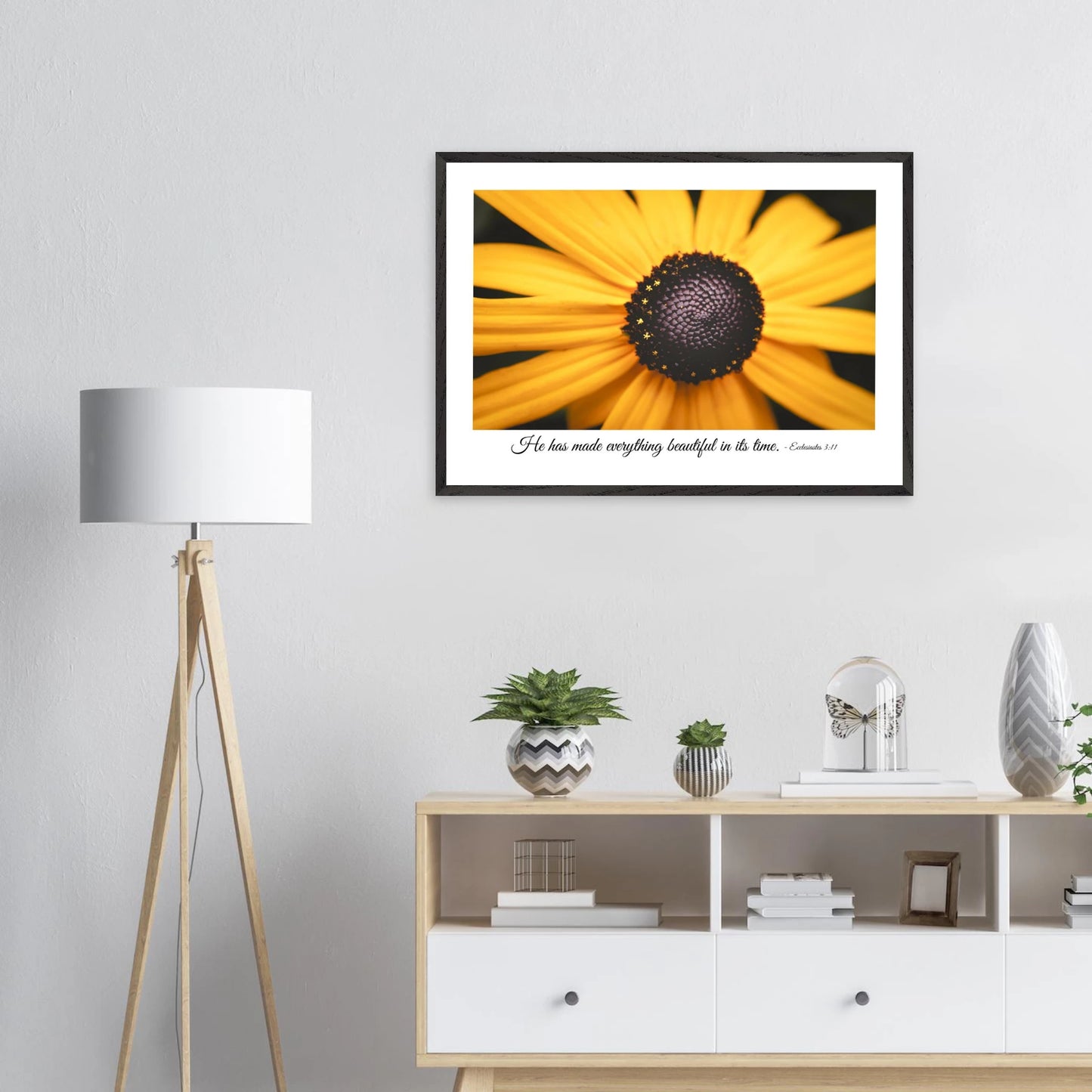 A framed art print of a close-up yellow flower with a quote from Ecclesiastes 3:11, 'He has made everything beautiful in its time.' displayed in a bright minimalist room with a small decors.