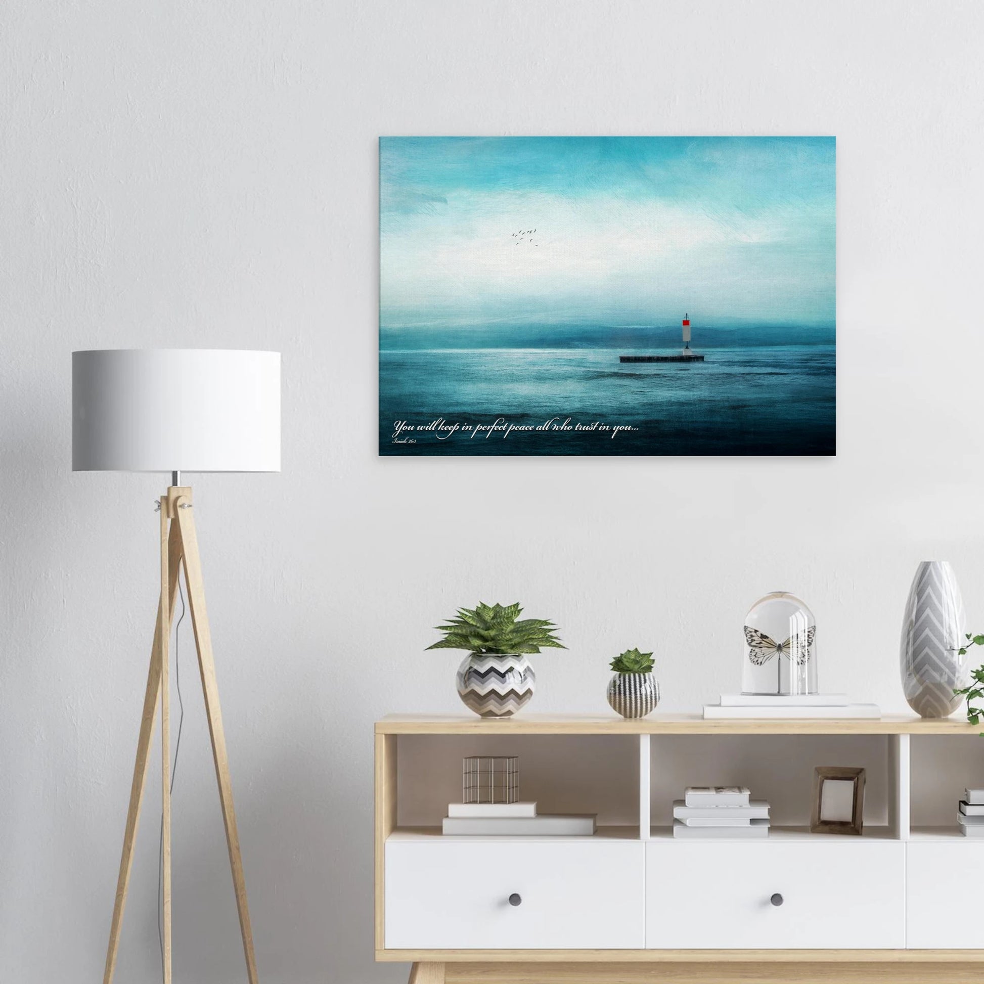 A wall art print featuring a lighthouse with a backdrop of the sea and sky, including a quote about perfect peace displayed in a bright minimalist room with lamp decor