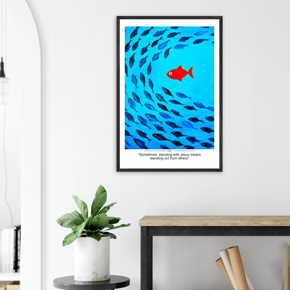 Framed Christian wall art featuring a red fish swimming in water with blue fish, and the text 'Sometimes standing with Jesus means standing out from others' as a quote displayed in a bright modern minimalist room and plant decor