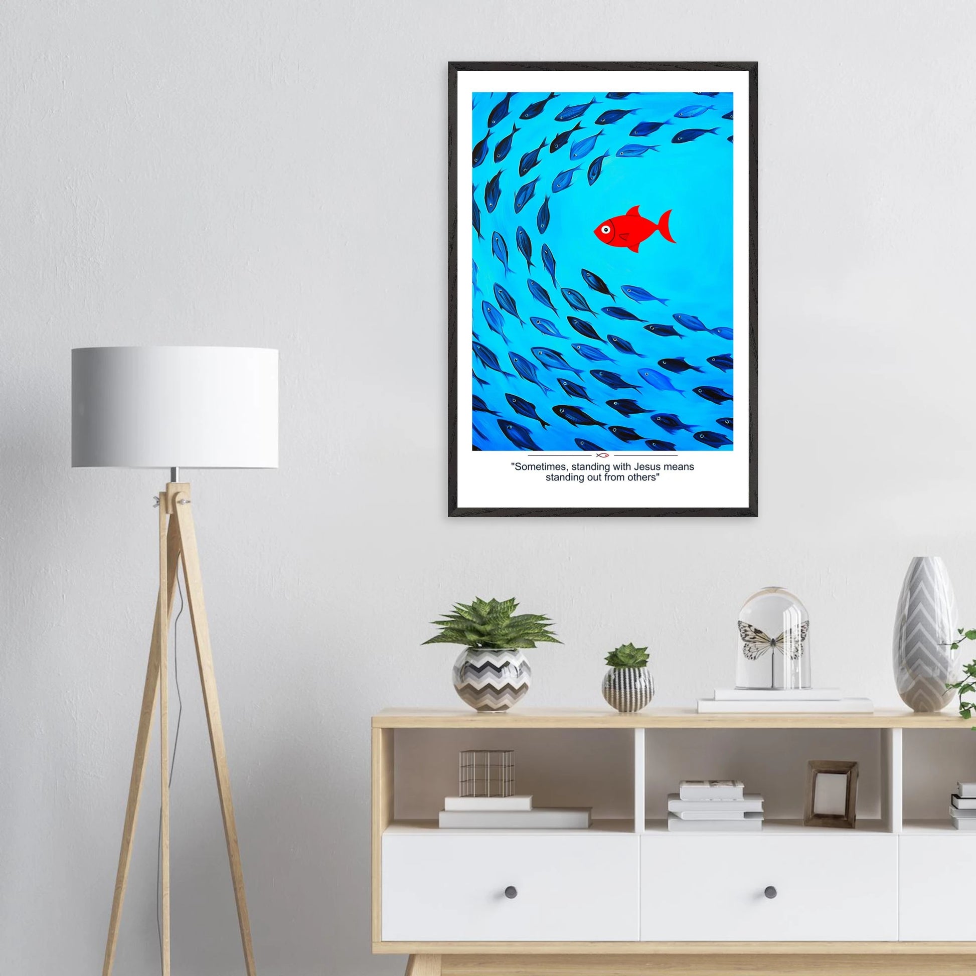 Framed Christian wall art featuring a red fish swimming in water with blue fish, and the text 'Sometimes standing with Jesus means standing out from others' as a quote displayed in a bright modern minimalist room