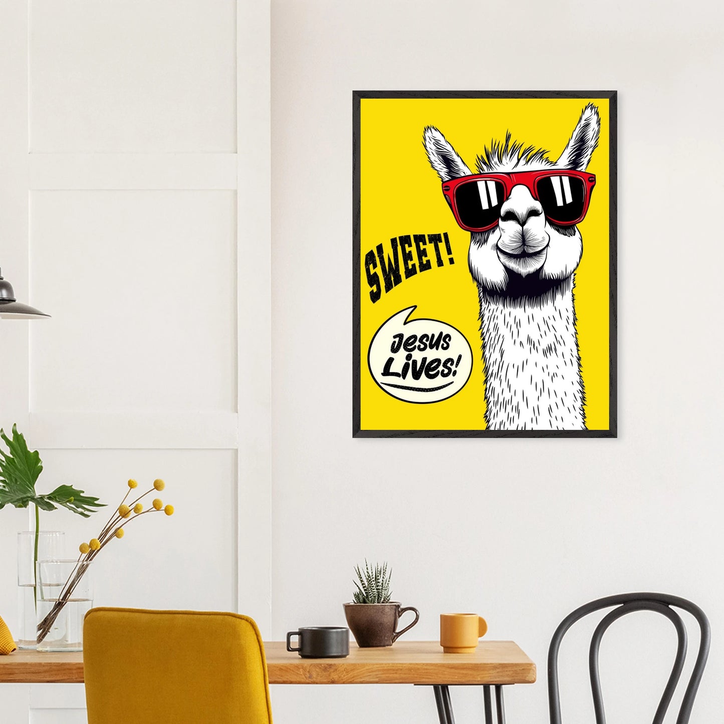 Framed Christian wall art featuring a white llama wearing red sunglasses on a bright yellow background with playful text reading SWEET and Jesus Lives, hanging above a wooden table in a modern dining space
