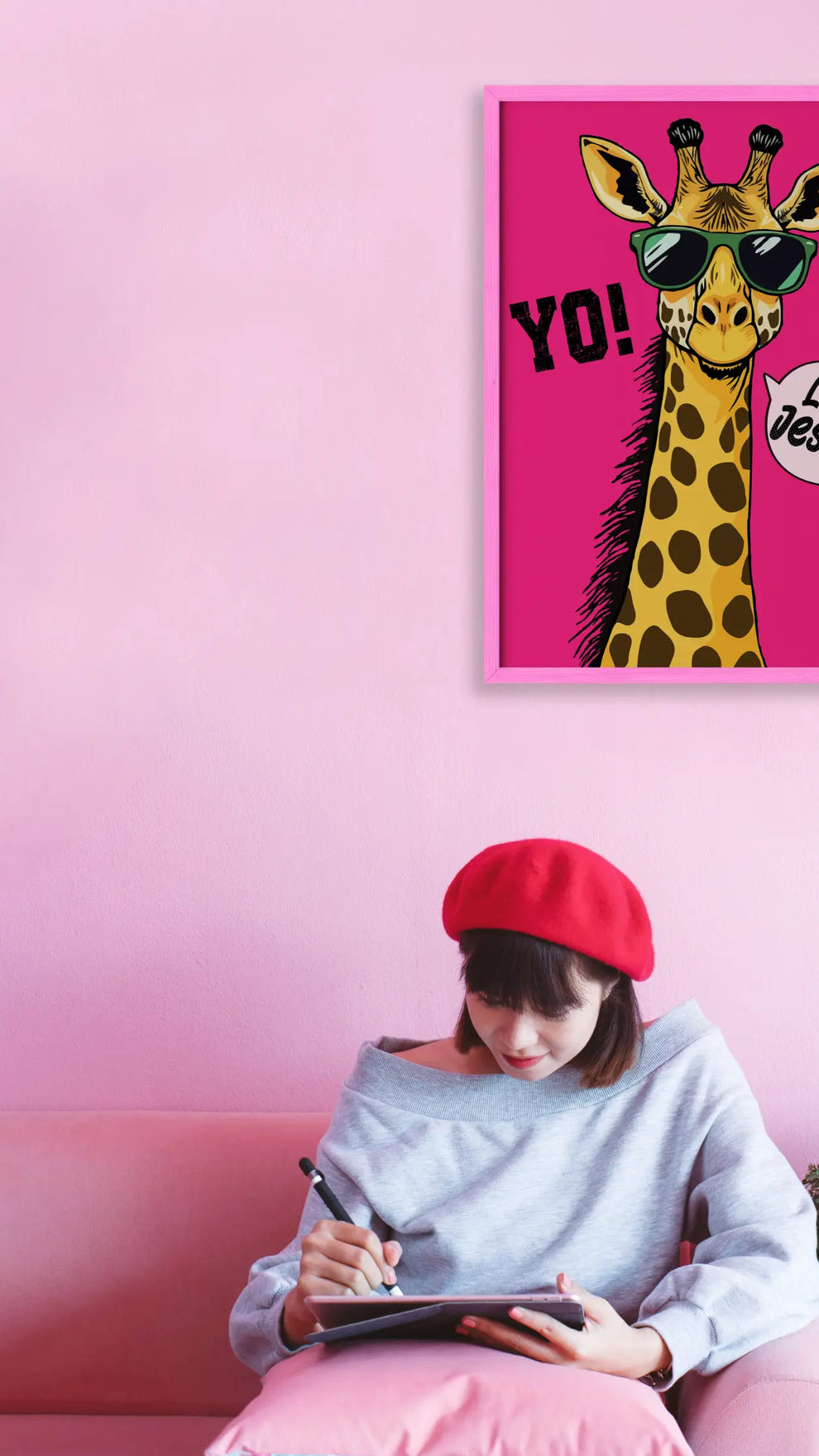 Woman sitting on a pink couch with a tablet, wearing a red beret, in front of a pink wall with a framed illustration of a giraffe.