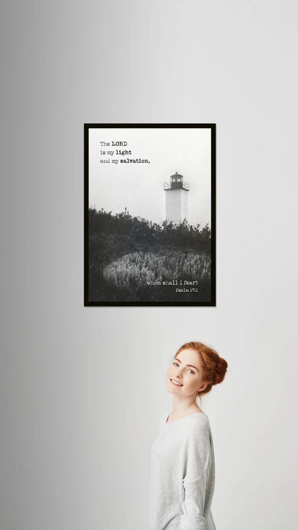 Framed black and white print of a lighthouse with text on a gray wall, with a person standing below.