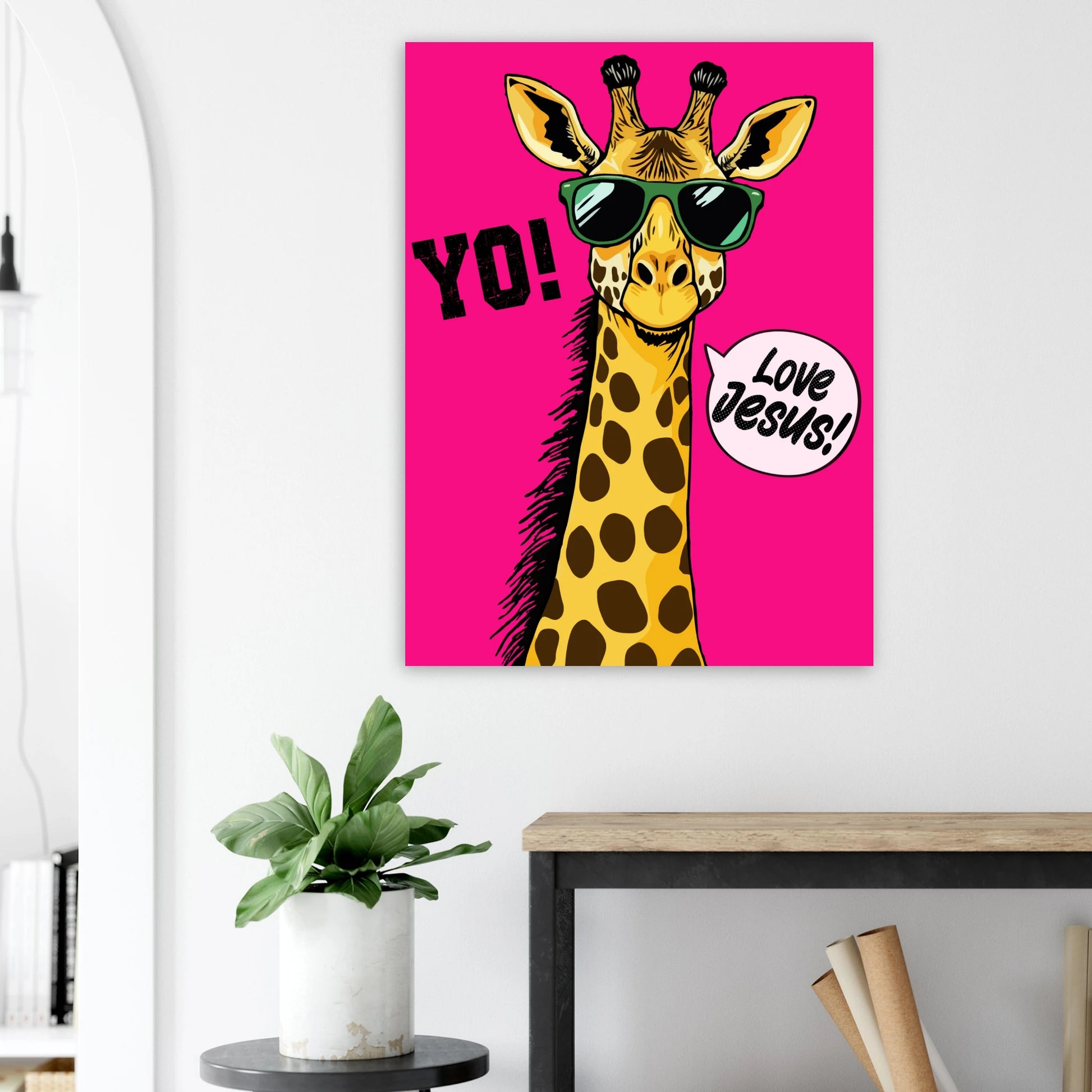 A colorful poster featuring a giraffe with sunglasses and a speech bubble that reads 'LOVE JESUS', set against a bold pink background displayed above a plant on a side table in a bright modern living room