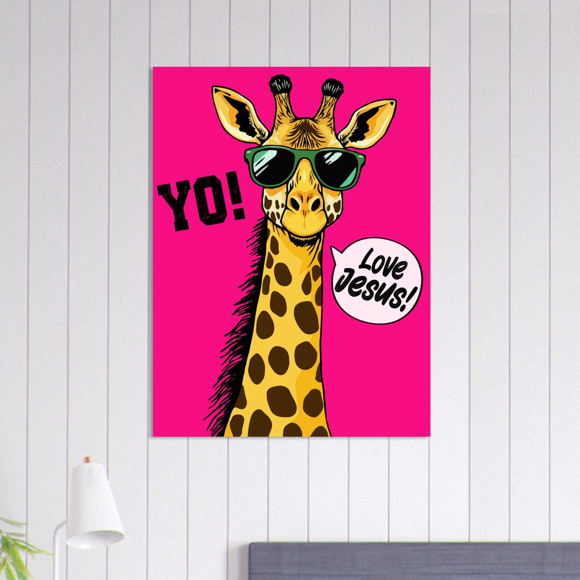 A colorful poster featuring a giraffe with sunglasses and a speech bubble that reads 'LOVE JESUS', set against a bold pink background displayed on a white paneled wall above a bed with a modern lamp nearby