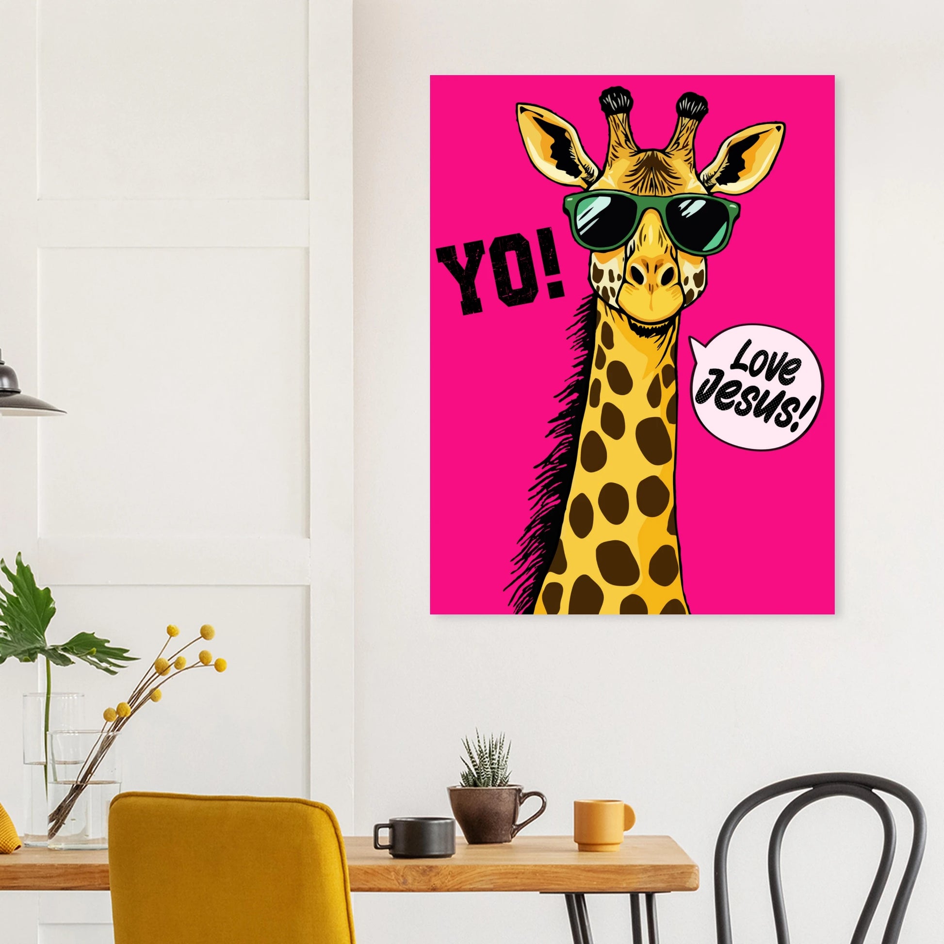 A colorful poster featuring a giraffe with sunglasses and a speech bubble that reads 'LOVE JESUS', set against a bold pink background hanging above a wooden table in a modern dining space