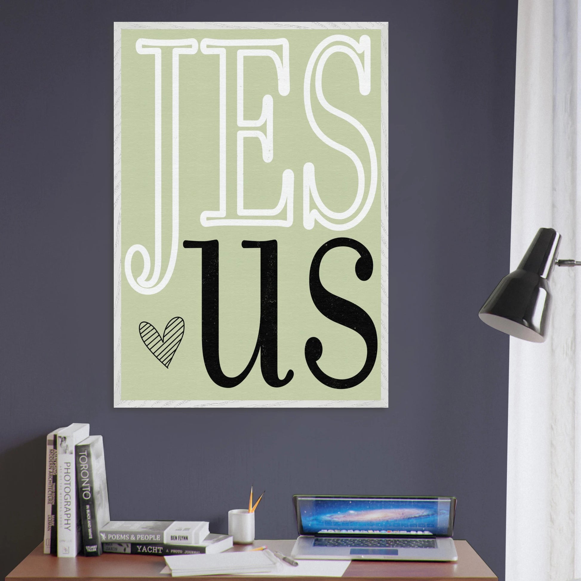Poster with 'JESUS' text on a wall above a desk with books and a laptop.