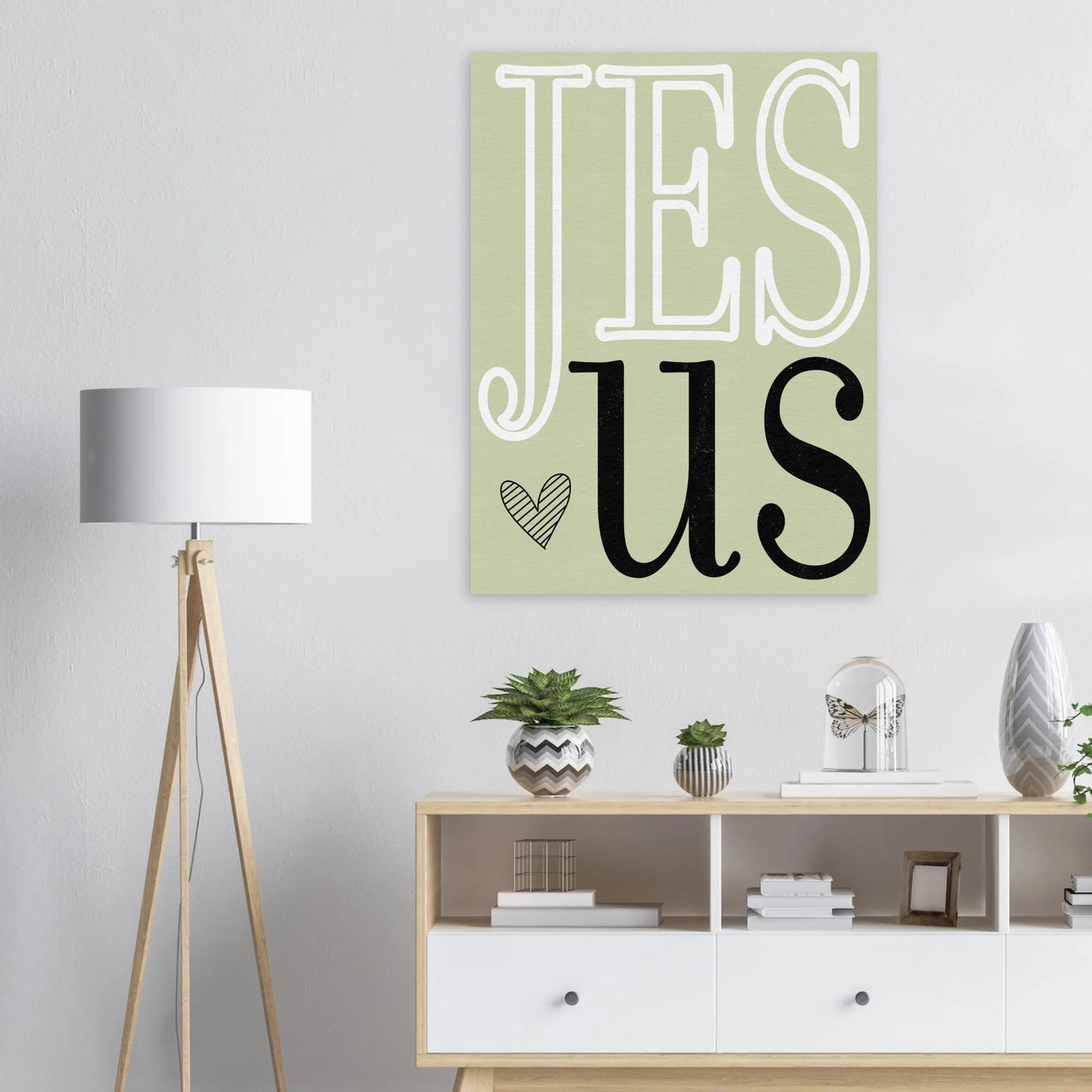 Decorative wall art with 'Jesus' on a light green background in a room with a lamp and shelves.