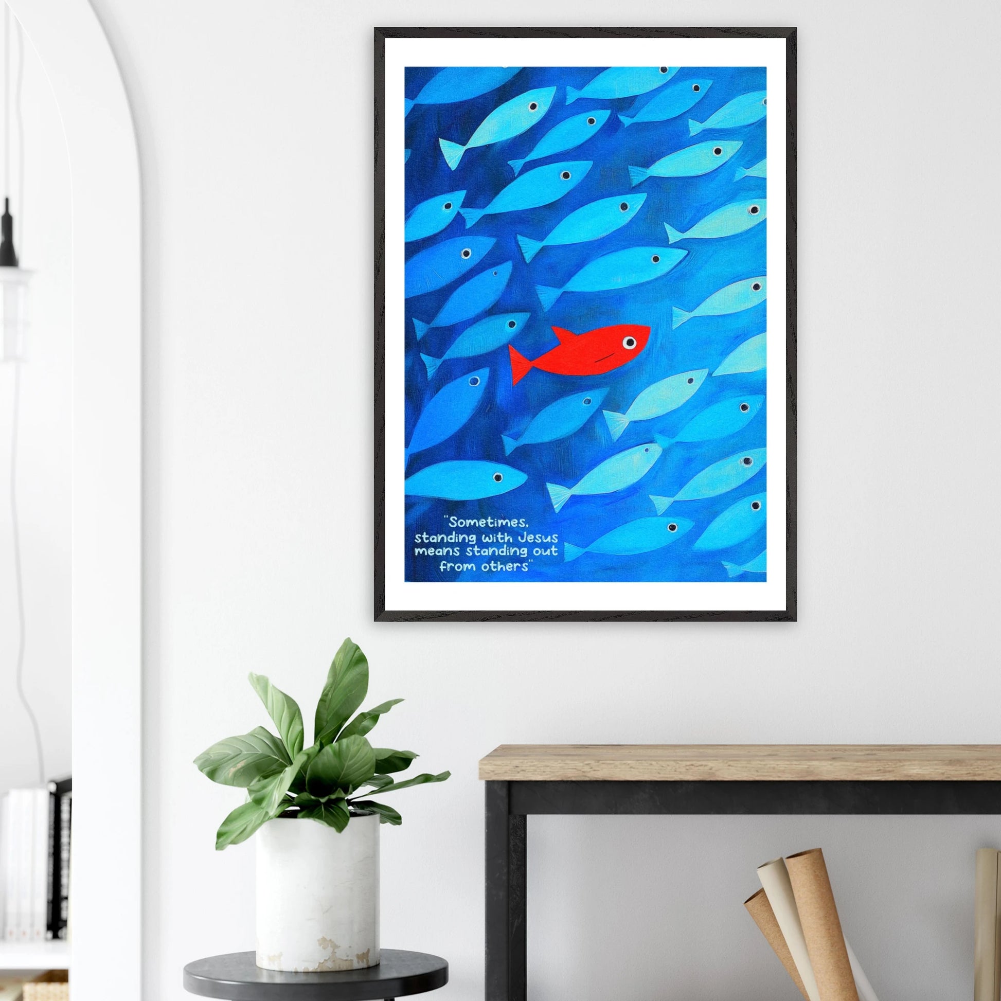 Framed wall art featuring a school of blue fish with a single red fish, and text that reads: 'Sometimes standing with Jesus means standing out from others.' displayed in a bright modern minimalist interior and plant decor