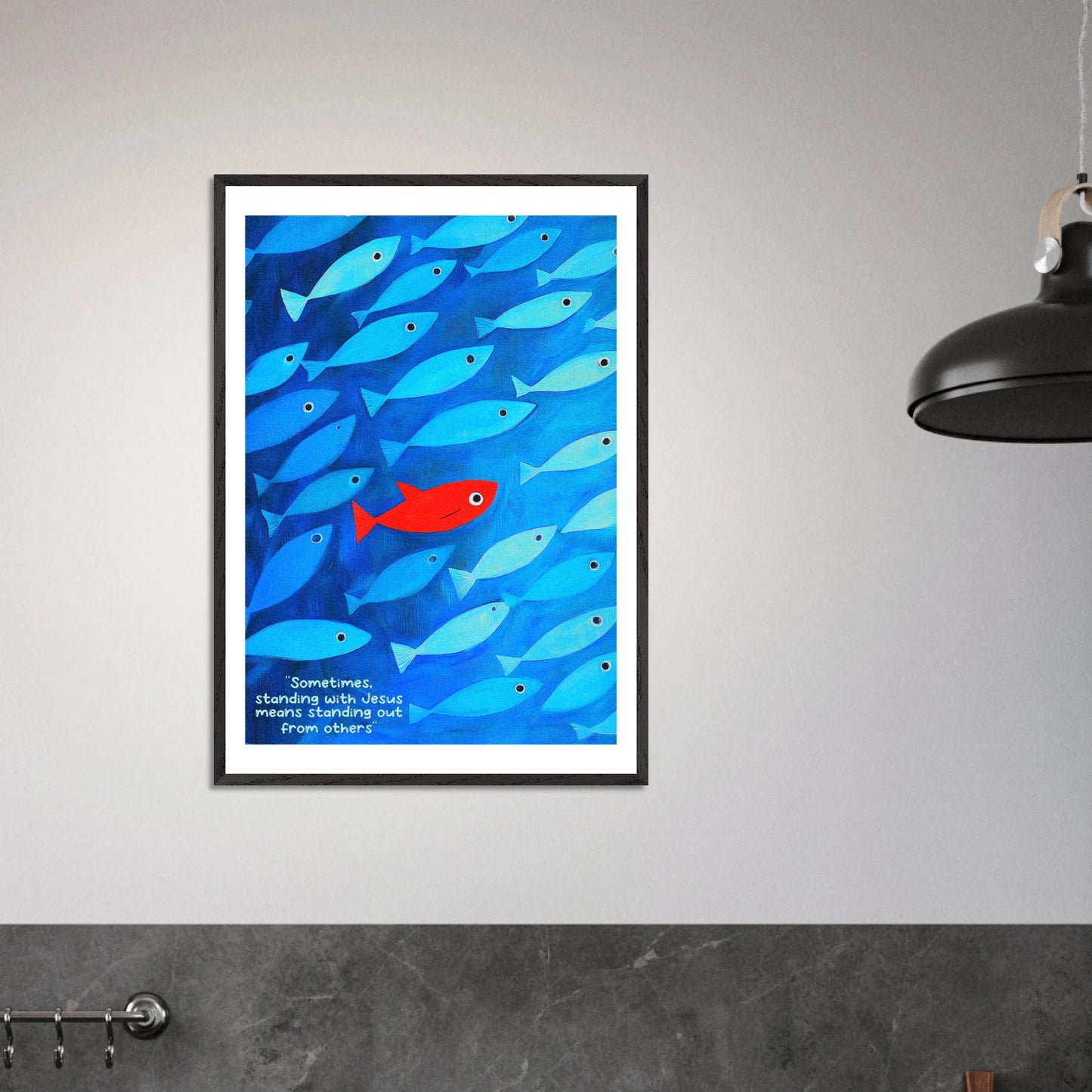 Framed wall art featuring a school of blue fish with a single red fish, and text that reads: 'Sometimes standing with Jesus means standing out from others.' displayed in a modern minimalist interior