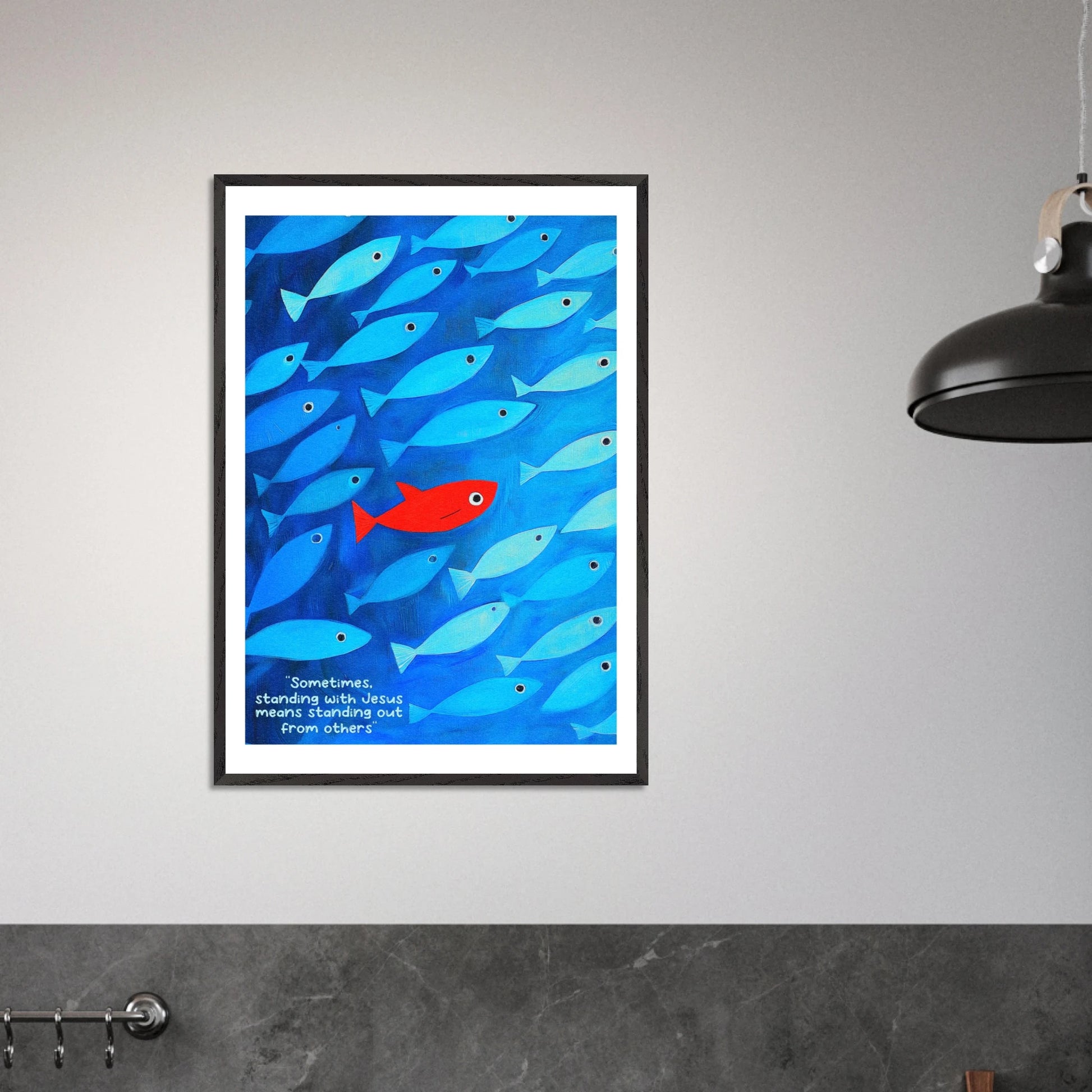Framed wall art featuring a school of blue fish with a single red fish, and text that reads: 'Sometimes standing with Jesus means standing out from others.' displayed in a modern minimalist interior