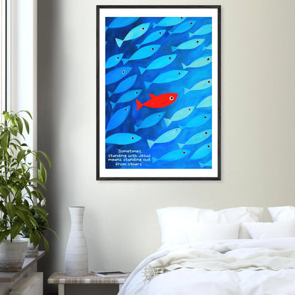 Framed wall art featuring a school of blue fish with a single red fish, and text that reads: 'Sometimes standing with Jesus means standing out from others.' displayed in a bright minimalist bedroom and plant decor