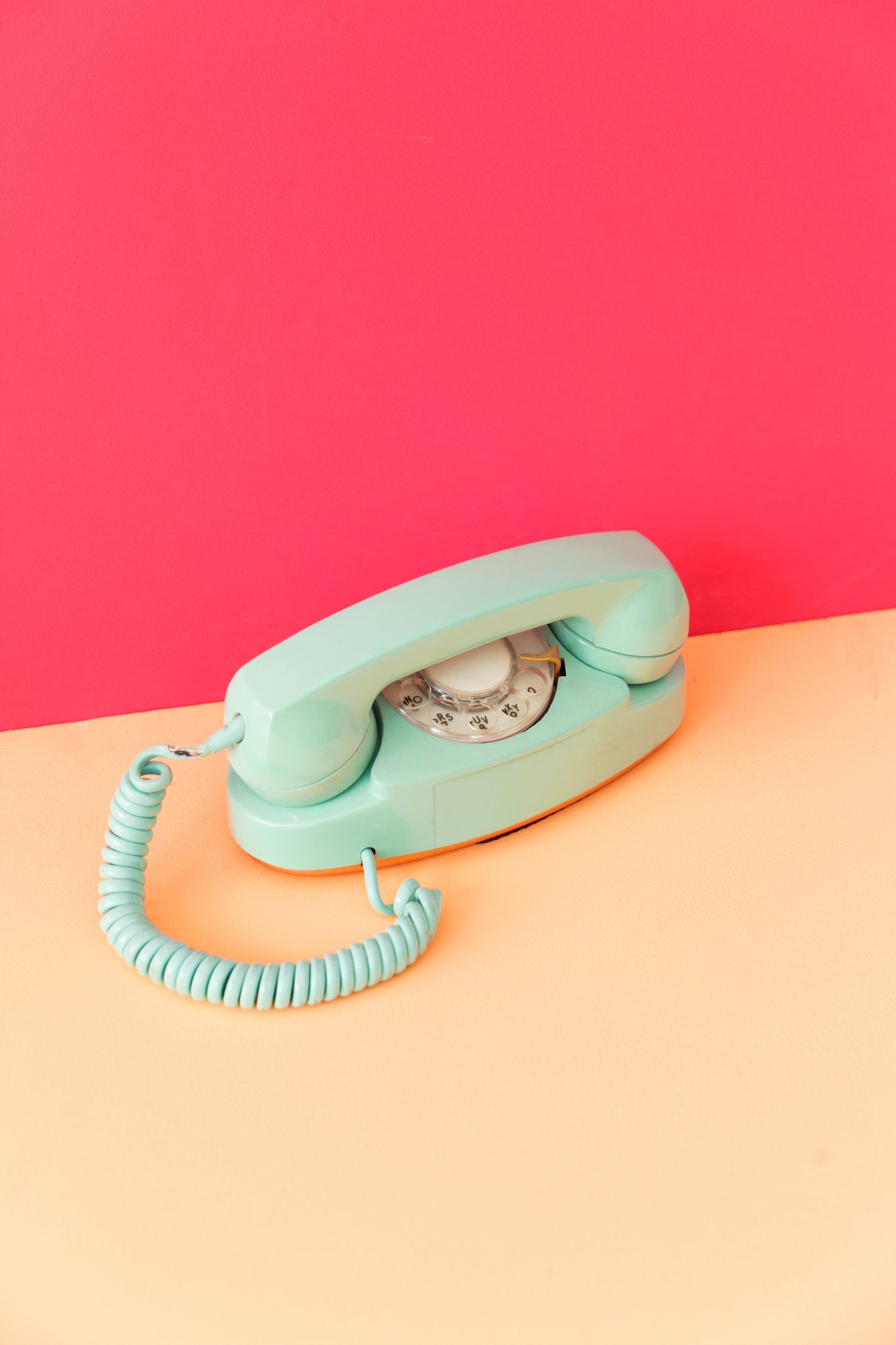 A mint-colored rotary telephone resting on a peach surface with a bold pink background behind it.