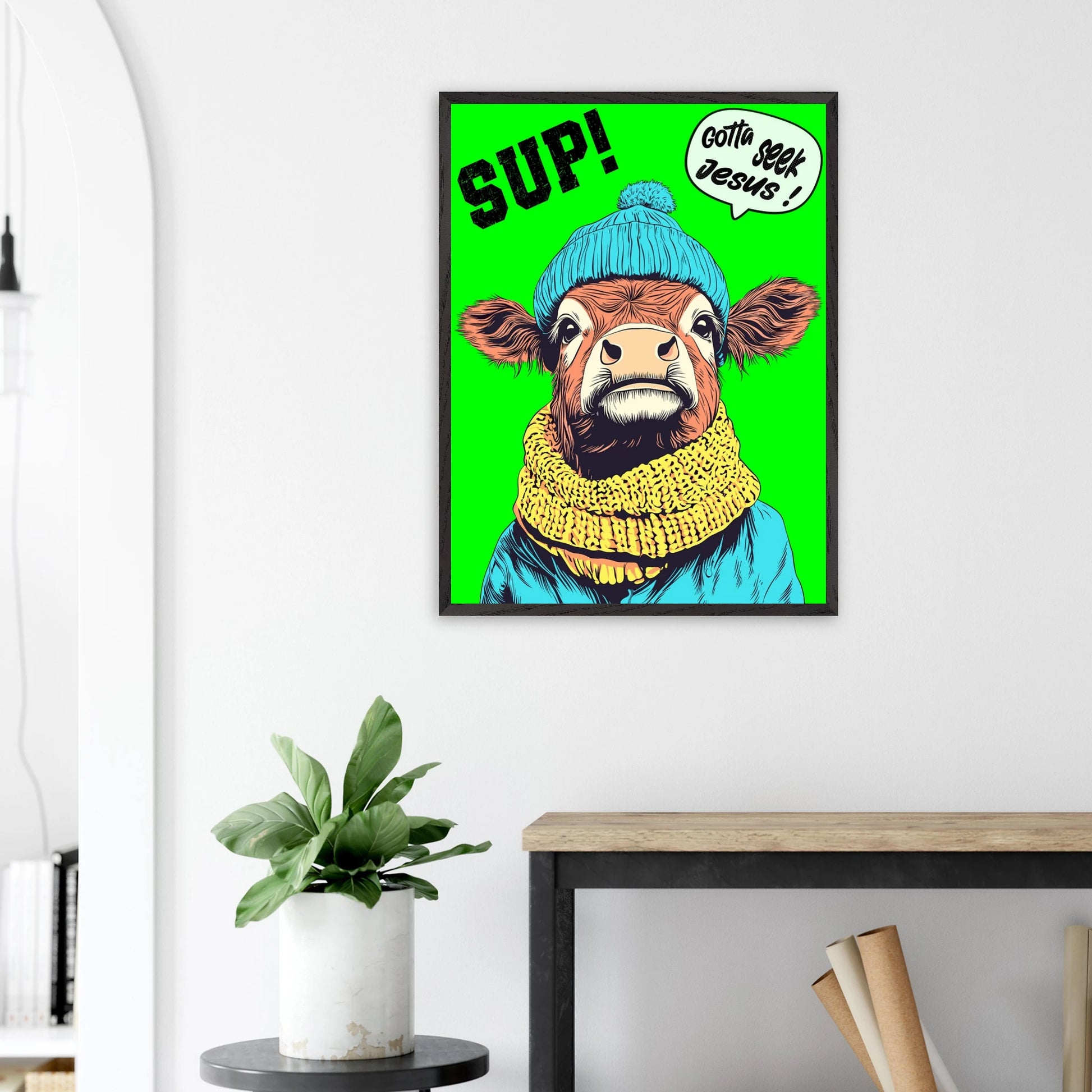 Playful Christian wall art featuring an illustrated cow wearing a blue beanie and yellow scarf on a bright green background, with SUP! and Gotta seek Jesus in a speech bubble, framed and displayed above a plant on a side table in a bright modern living room.