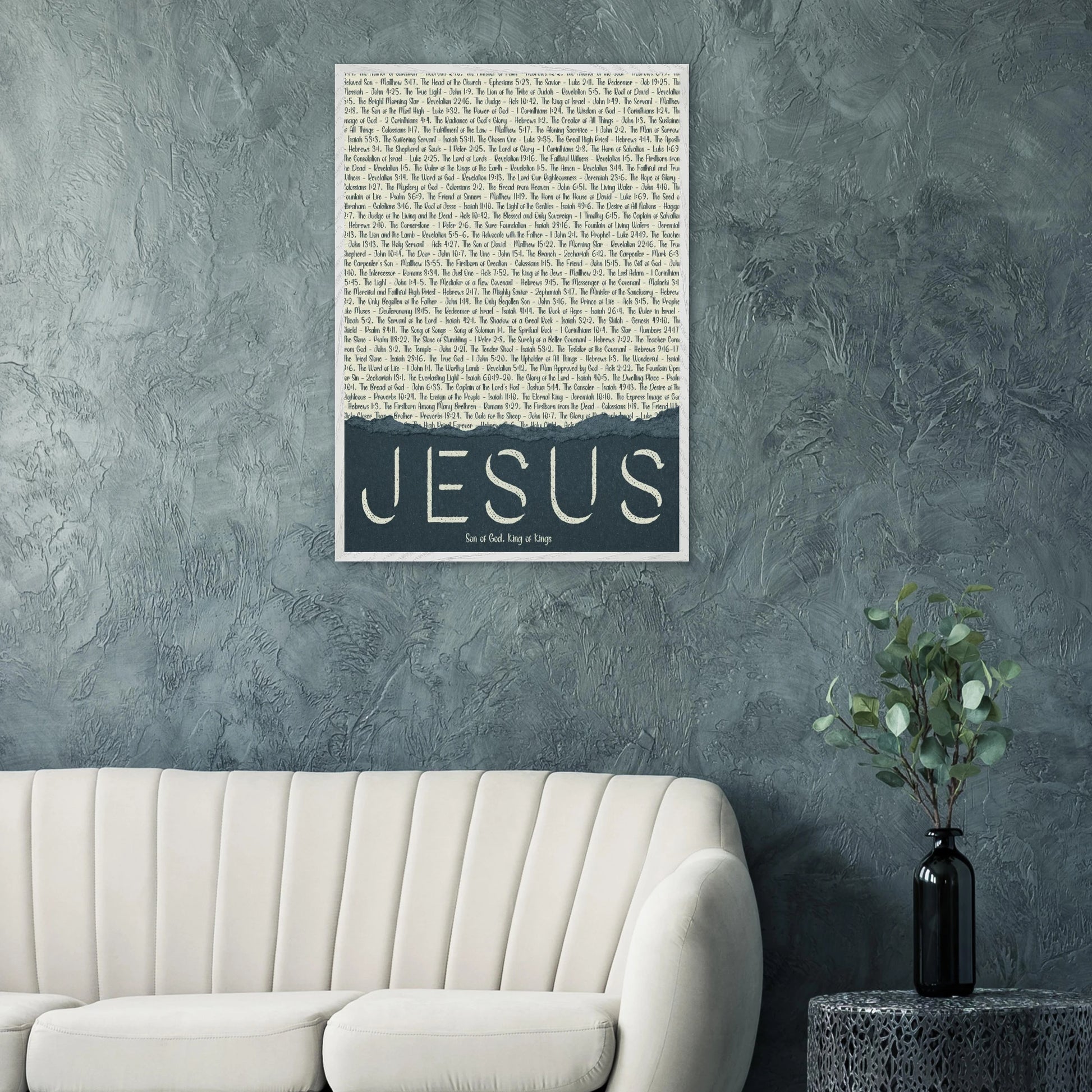 A framed wall art print with the word 'JESUS' in bold lettering, followed by a list of names and scriptures related to Jesus, against a white background hanging over a white couch in a living room