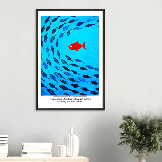 Framed Christian wall art featuring a red fish swimming in water with blue fish, and the text 'Sometimes standing with Jesus means standing out from others' as a quote displayed in a bright modern minimalist room and plant decor
