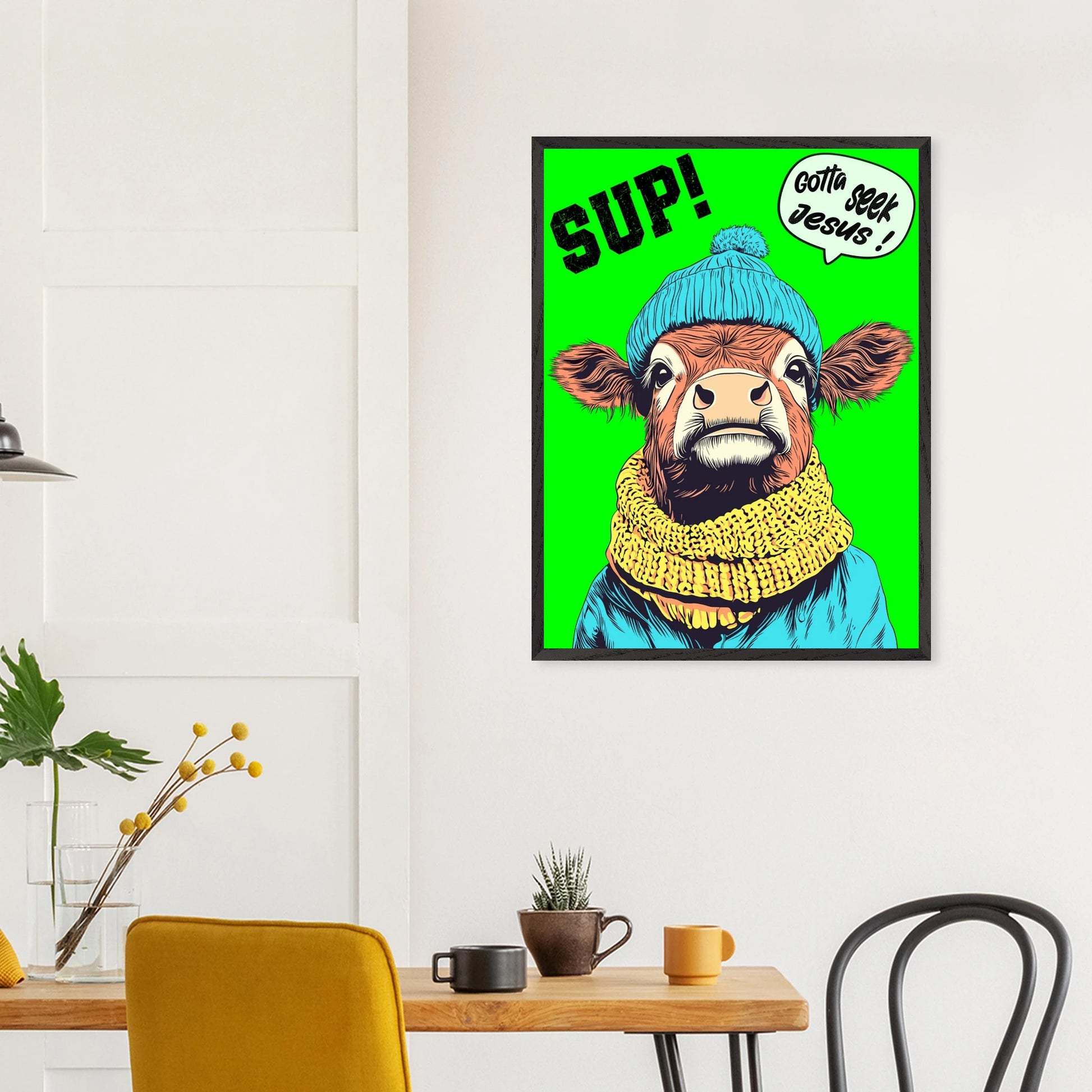 Playful Christian wall art featuring an illustrated cow wearing a blue beanie and yellow scarf on a bright green background, with SUP! and Gotta seek Jesus in a speech bubble, framed and hanging above a wooden table in a modern dining space
