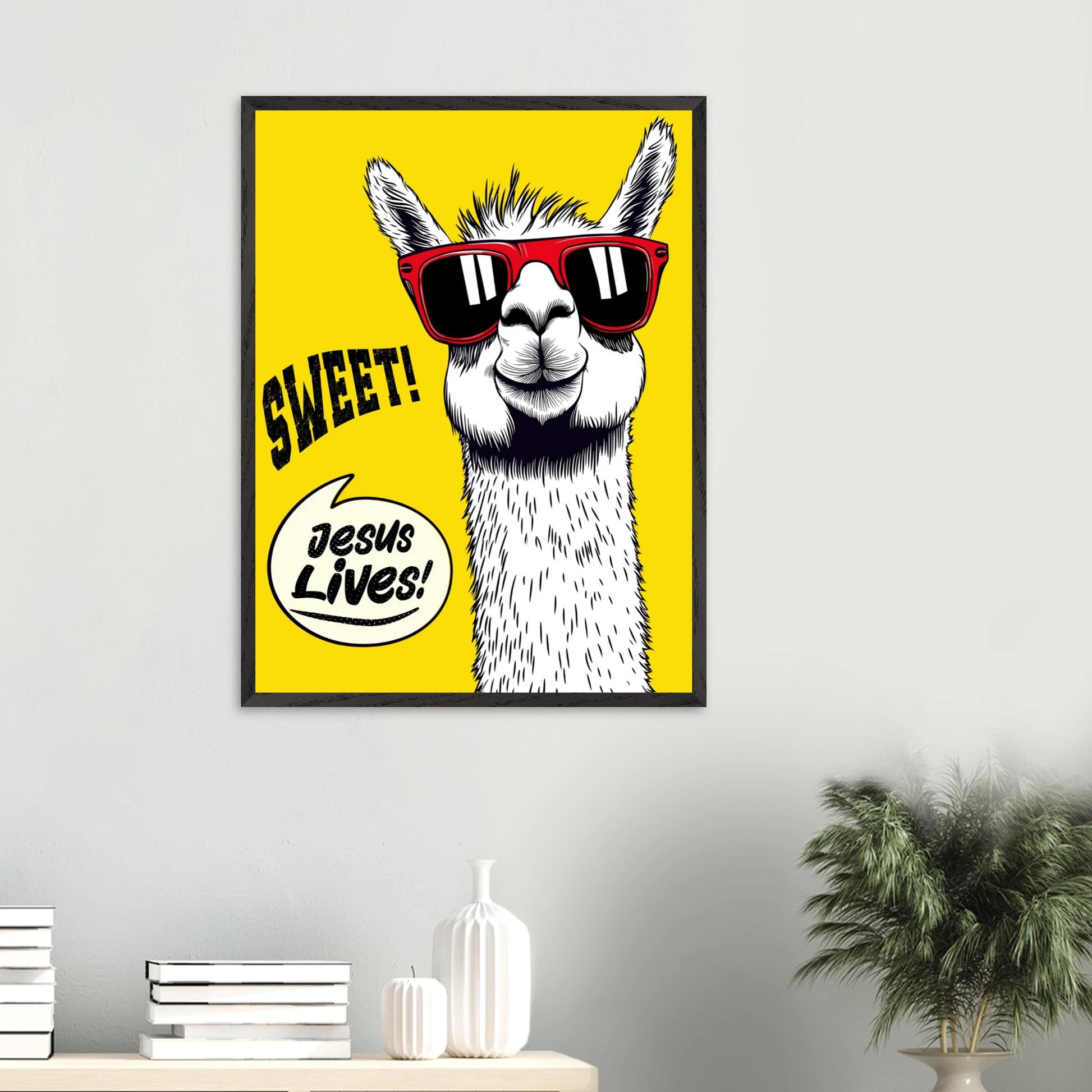 Framed Christian wall art featuring a white llama wearing red sunglasses on a bright yellow background with playful text reading SWEET and Jesus Lives, displayed on a minimalist white wall above a console with books, ceramic vases, and a green plant