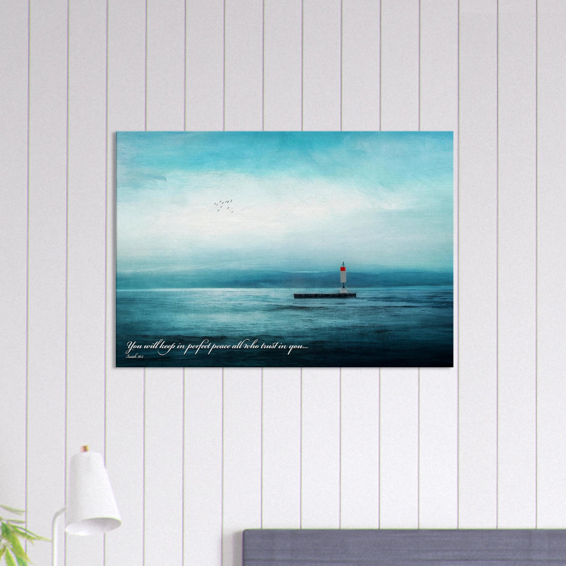 A wall art print featuring a lighthouse with a backdrop of the sea and sky, including a quote about perfect peace displayed in a bright minimalist room with lamp decor