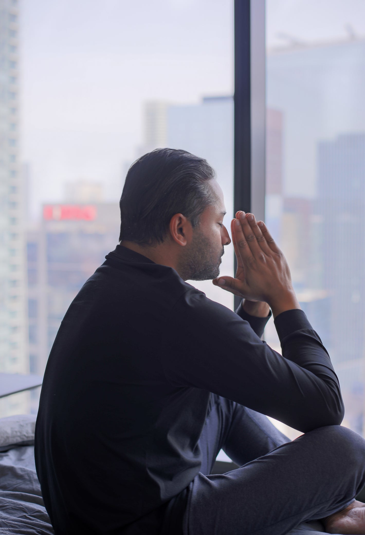 A person sits by a large window with hands pressed together, appearing deep in prayer or reflection.