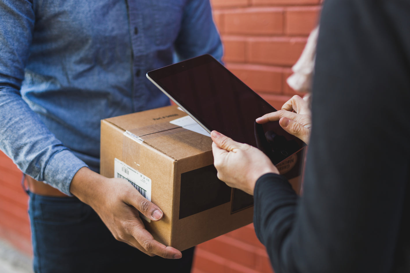A delivery person hands over a package while the recipient signs on a tablet.