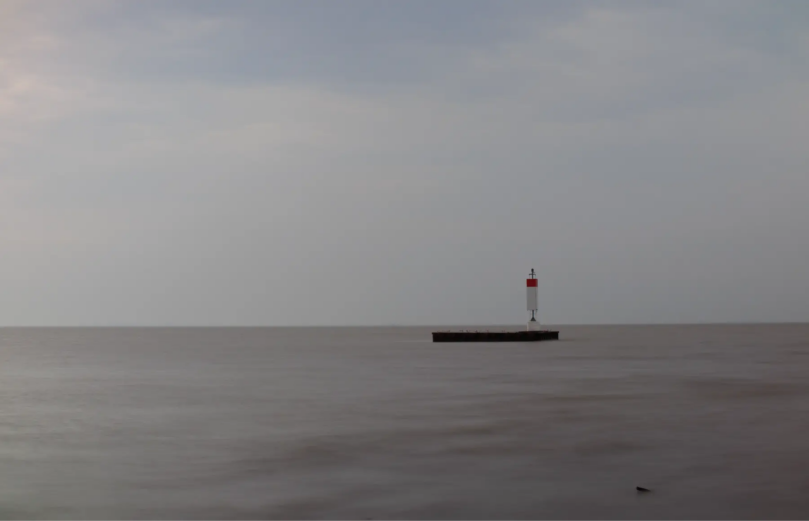 Lighthouse on a dock extending into a calm body of water under an overcast sky.